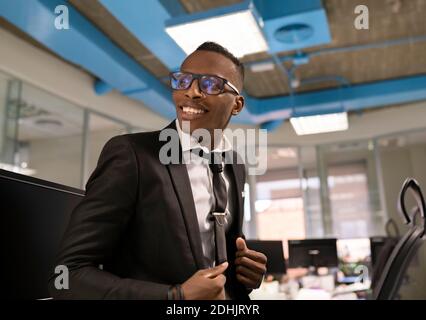 Low angle of positive young African American businessman dressed in elegant black suit with tie and eyeglasses working in contemporary workspace Stock Photo