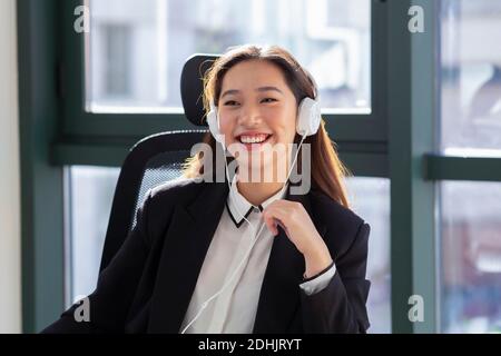 Happy young Asian female office employee in formal outfit and headphones laughing while having online conversation in modern office Stock Photo