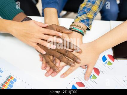 From above of crop anonymous multiracial colleagues in casual clothes gathering around table with charts and stacking hands together while working on Stock Photo