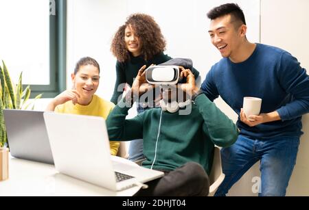 Unrecognizable black man with VR headset exploring virtual world while sitting at table with laptop surrounded by cheerful multiracial colleagues in m Stock Photo