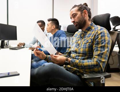 Side view of focused bearded male analyst manager in casual outfit examining paper with diagrams while working in modern workspace with colleagues Stock Photo