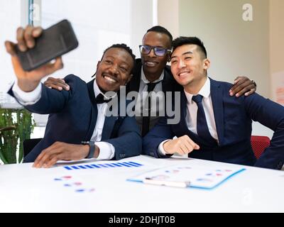 Group of smiling multiracial male business colleagues in formal suits gathering near table with diagrams and taking selfie on mobile phone during meet Stock Photo