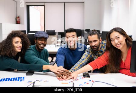 Young multiracial successful startup team gathering around table with diagrams and stacking hands together while discussing business strategy in moder Stock Photo