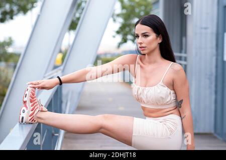 Side view of attractive young slim female in stylish sports outfit and sneakers doing stretching exercise for legs while standing near metal railing d Stock Photo
