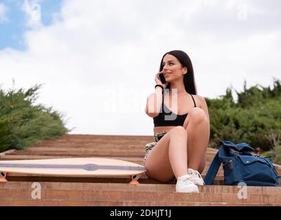 Low angle full length of smiling young fit female in activewear sitting on stairs near longboard and talking on mobile phone Stock Photo