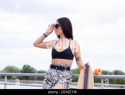 Confident young slim brunette in stylish activewear and trendy sunglasses holding longboard and looking away while standing near railing on embankment Stock Photo