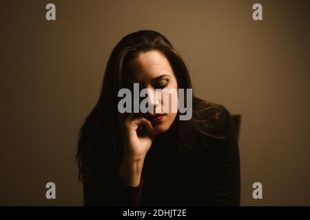 Unhappy pensive young female in black wear leaning on hand and thinking about personal problems while sitting against brown wall Stock Photo
