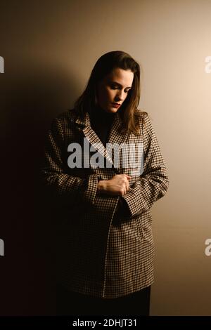 Sad lonely young brunette in trendy checkered coat looking down thoughtfully while standing against brown wall Stock Photo