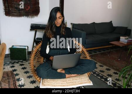 Serious Indian female freelancer sitting with crossed legs in wicker chair and typing on netbook while working online from home Stock Photo