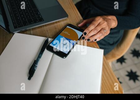 From above of crop anonymous female sitting at table with cup of coffee and analyzing diagram on smartphone while working on online project Stock Photo