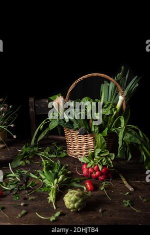 Wicker basket with various fresh green edible herbs placed on wooden table with bunch of radish and scattered leaves Stock Photo