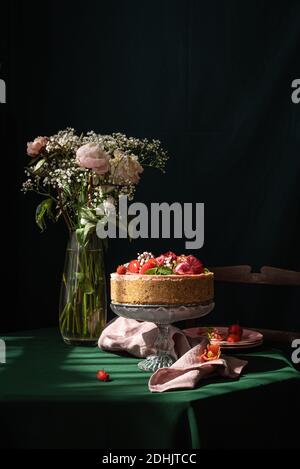 Still life composition with delicious homemade rustic cake decorated with fresh berries and flowers served on table near glass vase with delicate rose Stock Photo