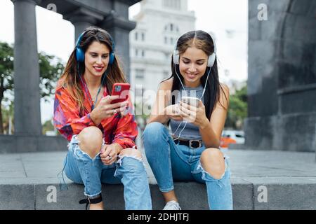 Cheerful hipster female students in trendy outfits surfing social networks on mobile phones and listening to music with headphones while sitting toget Stock Photo