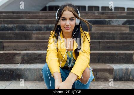Low angle of young female in trendy jacket and ripped jeans looking at camera while sitting on stairs near urban building and listening to music throu Stock Photo