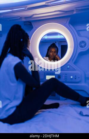 Side view of young African American female traveler looking at reflection in mirror while resting on bed in small room of capsule hotel with blue illu Stock Photo