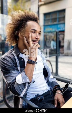 Pensive young black hipster male with curly Afro hair looking away thoughtfully while sitting at table in urban cafeteria Stock Photo