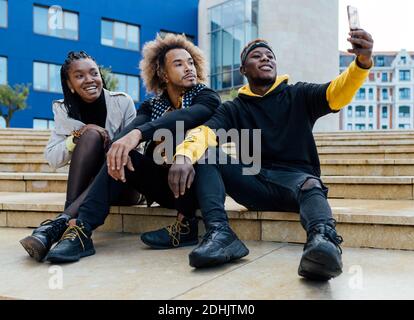 Full length of group of positive young African American students in casual clothes sitting on stairs on city street and taking selfie on mobile phone Stock Photo