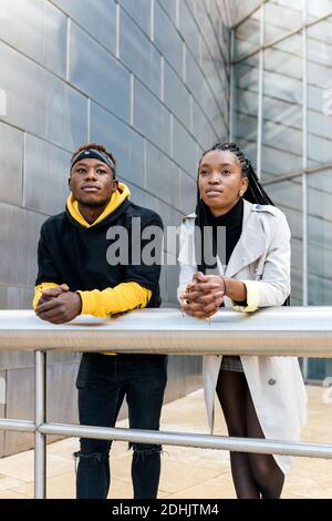 Young African American couple in trendy outfits leaning on railing and looking away thoughtfully while standing against contemporary urban building Stock Photo