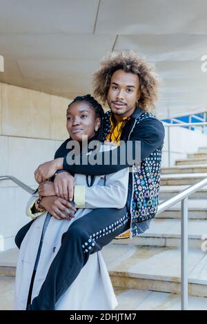 Happy young African American man embracing happy girlfriend while sitting on stairway rail near modern buildings during romantic date in city Stock Photo