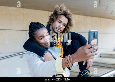Cheerful young African American couple hugging and taking selfie on smartphone while spending time together in city Stock Photo