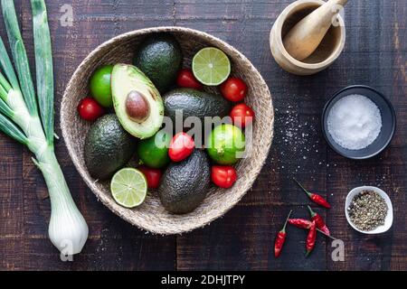 Top view bowl with fresh avocado and lime with tomatoes placed on wooden table with green onion and spices for traditional Mexican guacamole recipe Stock Photo