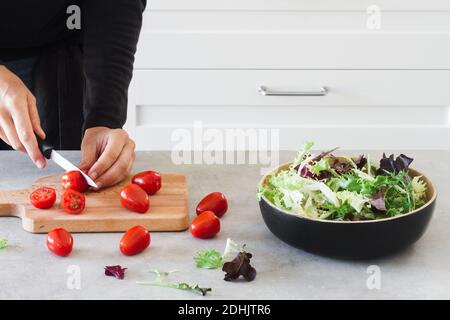 Faceless cook at gray countertop cutting cherry tomatoes on wooden board while making Caesar salad at home Stock Photo