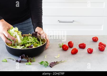 Faceless person mixing salad green leaves in bowl with hand at table with fresh tomatoes for Ceasar salad Stock Photo