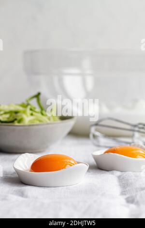 Ceramic bowls with separated raw egg yolks placed on table with fresh greens and mixer during breakfast preparation Stock Photo