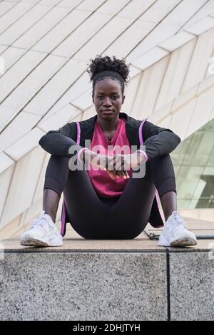 Determined African American female athlete in sportswear sitting on street and looking at camera Stock Photo