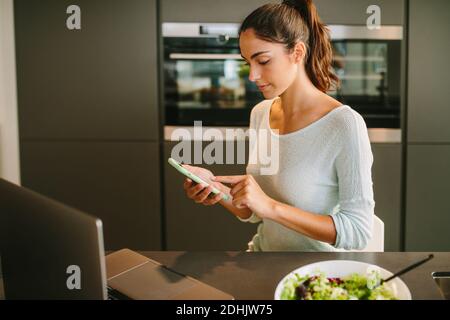 Young female remote worker in casual clothes checking messages on mobile phone while sitting at kitchen counter with laptop and bowl of salad Stock Photo