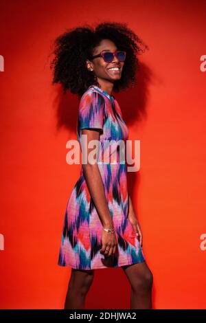 Happy black Woman with Afro hairstyle wearing vivid dress with sunglasses and laughing against red background Stock Photo