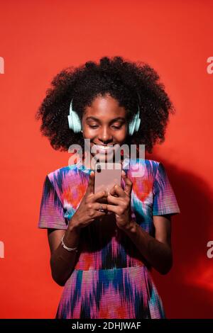 Bright Cuban model with Afro hairstyle listening to music with headphones and browsing smartphone on red background Stock Photo
