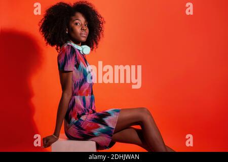 Full body side view of black Woman with Afro hairstyle wearing vivid dress with pink sneakers sitting on red background Stock Photo