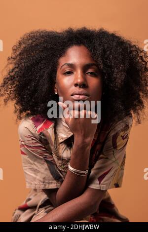 Serious African American model with Afro hairstyle leaning on hand and looking at camera on brown studio background Stock Photo