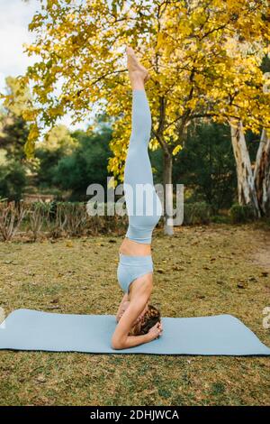 Side view of slender female balancing in Salamba Sirsasana on mat while doing yoga in park Stock Photo