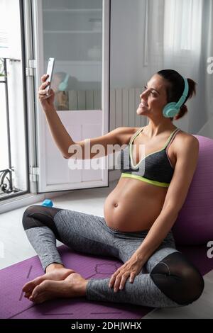 Side view of smiling pregnant female sitting on mat and taking selfie on smartphone while listening to music in headphones after practicing yoga Stock Photo