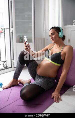 Pregnant female sitting on mat using smartphone while listening to music in headphones after practicing yoga Stock Photo