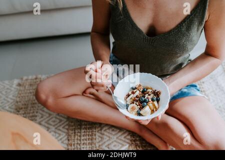 Cropped unrecognizable female with tasty bowl of healthy granola sitting on floor at home and having nourishing breakfast Stock Photo