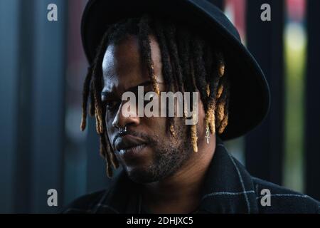 Informal bearded ethnic man with dreadlocks and in hat looking away standing on street Stock Photo