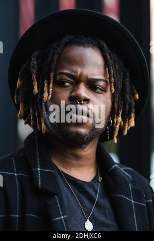 Informal bearded ethnic man with dreadlocks and in hat looking away standing on street Stock Photo