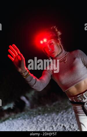 Young female traveler in red hiking headlamp keeping arm raised while exploring wild stony area at night Stock Photo