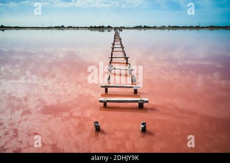 Drone view of old wooden walkway leading through pink water of calm lake towards distant coast Stock Photo