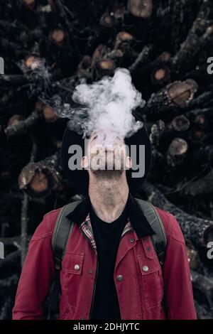 Male hipster in hat and red shirt exhaling cloud of smoke standing along with eyes closed against pile of logs in nature Stock Photo