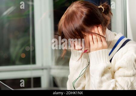 a young pretty girl in headphones sitting at a laptop learns a foreign language on the windowsill at the window Stock Photo