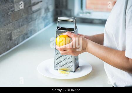 Female hands grating fresh whole lemon on metal hand grater, making zest on her kitchen table. Yellow and gray trendy colors of the year 2021 Stock Photo
