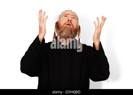 Praying bearded Jewish man with sidelocks in white kippah raising his hands to heaven. Studio portrait of hasid wearing black coat isolated on white Stock Photo