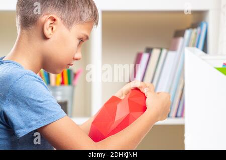 Boy looking video tutorial and making red polygonal paper heart. Education, learning, paper craft, entertainment at home Stock Photo