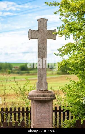 Zadni Kopanina, Czech Republic - August 23 2020: A stone cross standing inside a wooden fence in field with green tree. Colorful background. Sunny day. Stock Photo