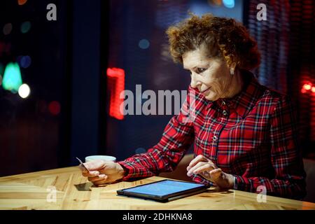 Mature woman makes online purchases using tablet computer in cafe. Older female pays for online payment with credit card. Modern senior uses plastic c Stock Photo