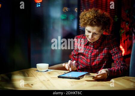 Senior woman ordering Christmas gifts online on digital tablet in coffee shop. Mature buyer holding tablet pc and credit card. Elderly woman shopping Stock Photo